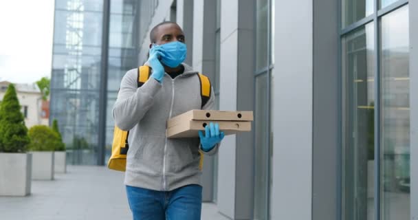 身穿手套和医疗面罩的年轻的非洲裔美国男性送货工人在街上走来走去，一边拿着披萨盒一边打电话。男性信使在手机上讲话. — 图库视频影像
