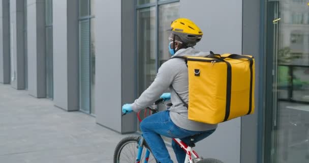 Arrière d'un coursier afro-américain avec un gros sac au dos en casque, masque médical et gants à vélo dans la rue et livrant de la nourriture ou des colis. Vue arrière sur livreur cycliste . — Video