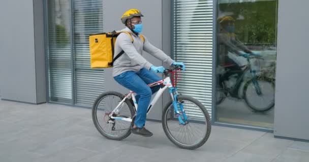 Joven guapo afroamericano en casco, guantes y máscara médica montando en bicicleta y trabajando como repartidor. Entrega del concepto de bicicleta en cuarentena . — Vídeos de Stock