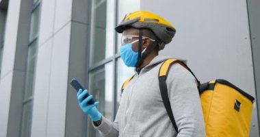 Tıbbi maskeli Afro-Amerikalı teslimatçı, eldivenler ve çarşaf dışarıda bisiklet başında duruyor ve akıllı telefondan mesaj yolluyor. Bisikletli kurye caddede cep telefonunu dinliyor. Konsept sunma.