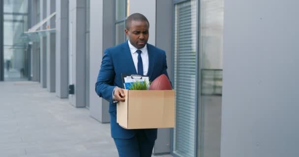Smutny młody Afroamerykanin w garniturze i krawacie chodzący ulicą, niosący pudełko z rzeczami po zwolnieniu z pracy. Bezrobotny pracownik biurowy spacerujący na zewnątrz. Pożar podczas kryzysu zamknięcia. — Wideo stockowe