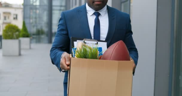 Trabajador de oficina afroamericano deprimido saliendo del edificio del centro de negocios con caja de cosas personales. El hombre de negocios perdió el trabajo. Hombre despedido caminando al aire libre. Desempleado deprimido. Desempleo. — Vídeos de Stock