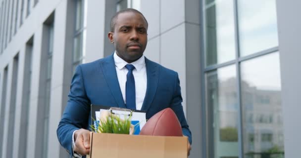 Retrato de un joven empresario afroamericano triste parado al aire libre con caja de cosas en el centro de negocios y mirando a la cámara. Trabajador de oficina perdió el trabajo. Tasa de desempleo en pandemia. Hombre despedido . — Vídeos de Stock