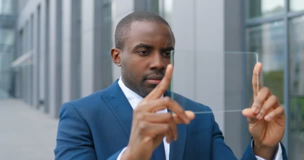 Joven hombre de negocios afroamericano en traje y corbata de pie en la calle y sosteniendo un pedazo de vidrio en el que ver algo como en la pantalla digital. Tecnología del futuro. Dispositivo transparente . — Vídeos de Stock