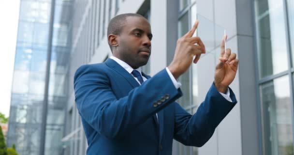 Jeune homme d'affaires afro-américain en costume et cravate debout à la rue et tenant un écran transparent en verre sur lequel regarder quelque chose comme sur l'écran numérique. Dispositif technologique futur . — Video