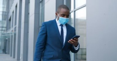 Tıbbi maskeli Afro-Amerikalı işadamı sokakta yürüyor ve cep telefonundan mesaj yolluyor. Solunum korumalı erkek yaya dışarıda dolaşıyor ve akıllı telefonları dinliyor ya da kaydırıyor..