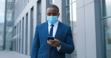 Tıbbi maskeli Afro-Amerikalı işadamı caddede duruyor ve cep telefonundan mesaj yolluyor. Solunum korumalı erkek yaya dışarıda dolaşıyor ve akıllı telefonları dinliyor ya da kaydırıyor..