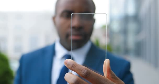 Primer plano del joven empresario afroamericano en traje y corbata sosteniendo pantalla transparente de vidrio, viendo algo. Pantalla del teléfono. Al aire libre. Futuro dispositivo de tecnología. Tecnología futurista. Borrosa. . — Vídeo de stock