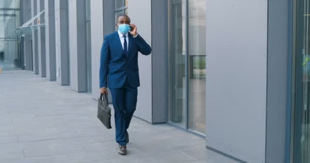 Afroamerikansk ung affärsman i medicinsk mask går på gatan och pratar i mobiltelefon. Manlig fotgängare i andningsskydd promenader utomhus och talar på mobiltelefon. Samtal. — Stockvideo