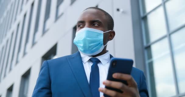 Primo piano di uomo d'affari afroamericano in maschera medica messaggio di sms sul telefono cellulare. Maschio in tuta, cravatta e protezione respiratoria in piedi all'aperto e toccando o scorrendo sullo smartphone . — Video Stock