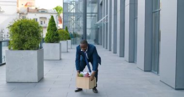 Afro-Amerikalı mutlu iş adamı iş merkezinden ayrılıp dans etmek gibi kutularla sokakta yürüyor. Erkek ofis çalışanı işi bırakıp kutlama yapıyor. Ateşli neşeli adam.