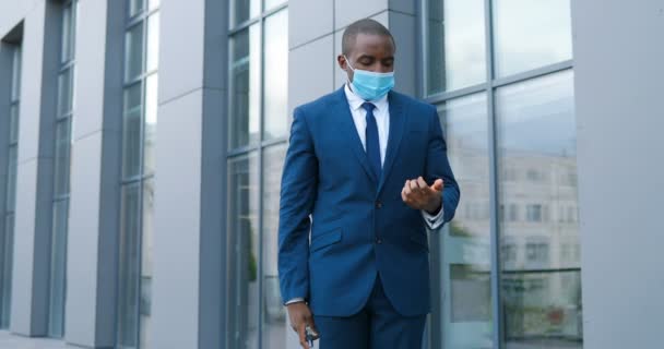 Joven empresario afroamericano enmáscara médica, traje y corbata rociando desinfectante en las manos y limpiándolas de gérmenes en la calle. Hombre durante la pandemia de coronavirus desinfectando la piel. Limpio y seguro . — Vídeo de stock