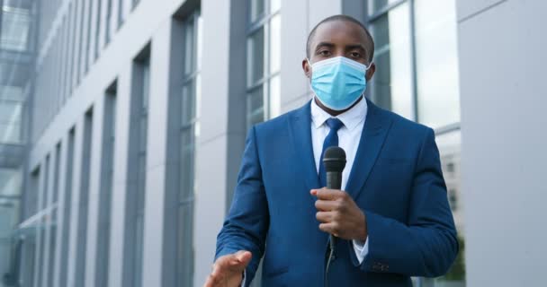 Porträtt av ung afroamerikansk stilig manlig journalist i medicinsk mask talar med mikrofon för nyhetsepisod utomhus. Ett pandemiskt koncept. Man korrespondent i kostym och slips och med mick. — Stockvideo