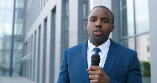 Portret van jonge Afro-Amerikaanse knappe mannelijke journalist in gesprek met microfoon voor nieuws aflevering buiten. Pandemisch concept. Close-up van de man correspondent in pak en das en met mic. — Stockvideo