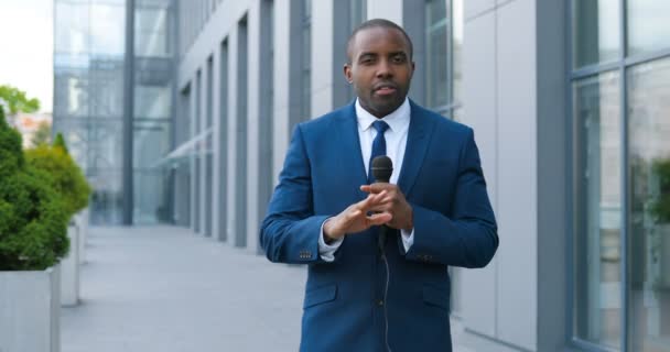 Portretfoto van jonge Afro-Amerikaanse knappe mannelijke journalist die praat met microfoon voor nieuws aflevering buiten. Pandemisch concept. Man correspondent in pak en das en met microfoon. — Stockvideo