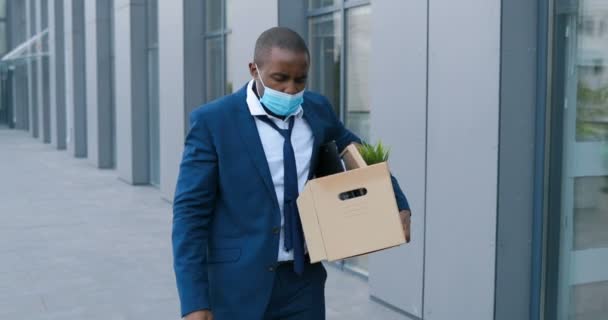 Afroamericano enojado empresario molesto en máscara médica caminando al aire libre con caja de cosas. Trabajador de oficina perdió el trabajo. Tasa de desempleo que crece con el brote de virus. Hombre despedido en la desesperación . — Vídeos de Stock