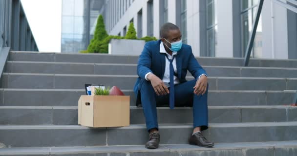 Despedido trabajador de oficina afroamericano en máscara médica sentado en las escaleras en la depresión con caja de cosas. El hombre de negocios desempleado perdió su negocio. Concepto ansioso. Hombre sin trabajo en la desesperación . — Vídeos de Stock