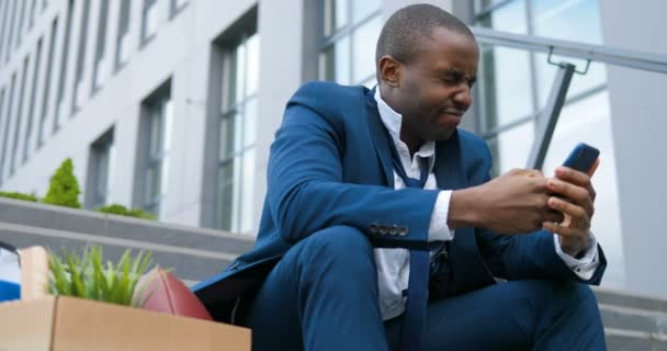 Afro-Amerikaanse zakenman zit buiten op de trap met een doos met persoonlijke kantoorspullen. Boos wanhopige man verloren werkplek en sms-bericht op smartphone. Een pandemische economische crisis. Ontslagen man. — Stockvideo