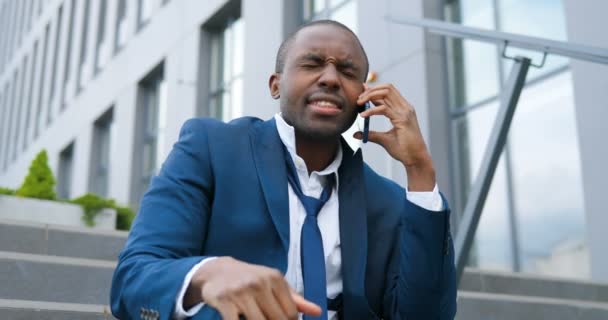 Nuori afrikkalainen mies työtön toimistotyöntekijä puvussa ja solmio istuu portaissa ulkona puhumassa kännykkään. Erotettu mies puhuu puhelimessa kadulla. Puhelinkeskustelu. — kuvapankkivideo