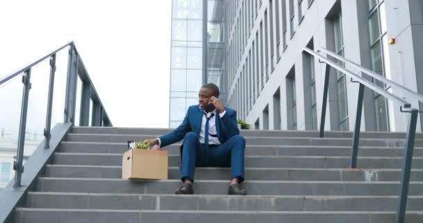 Молодий афроамериканець, безробітний офісний працівник у костюмі і краватка, сидить на сходах на відкритому повітрі. Звільнений чоловік розмовляє по телефону і дивиться на речі в коробці. Мобільна розмова. Говорить по телефону.. — стокове відео