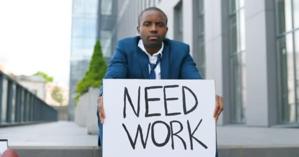 Afro-Américain jeune homme triste et déprimé dans le désespoir assis sur les escaliers dans la ville, montrant affiche Besoin de travail à la caméra. Homme sans travail regardant anxieux dans la dépression. Chômage au lock-out . — Video