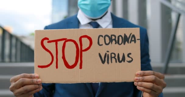 Закрыть картонный стол словами Stop Coronavirus в руках афроамериканского молодого человека в медицинской маске. Мужской активист. Акция протеста на улице . — стоковое видео