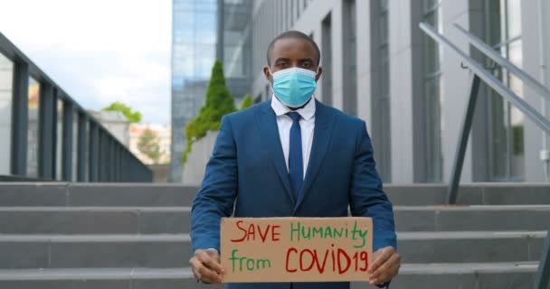 Портрет афроамериканского активиста-мужчины в медицинской маске с плакатом "Спаси человечество от коронавируса" на одинокой демонстрации в городе. Единственный протест снаружи. Концепция активизма. Человек, протестующий в одиночку — стоковое видео