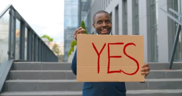 Retrato de un hombre afroamericano sosteniendo un cartel Sí y haciendo un gesto de dinero en una manifestación o protesta. Una sola protesta al aire libre. Concepto de activismo. Hombre alegre mostrando mesa con la palabra sí . — Vídeos de Stock