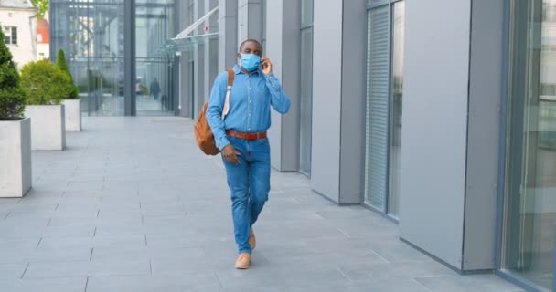 Афроамериканець у медичній масці ходить по вулиці і розмовляє по мобільному телефону. Чоловічий пішохід під час захисту дихальних шляхів крокує надворі і розмовляє по мобільному телефону. Розмова. — стокове відео