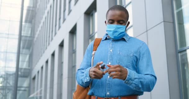 Il giovane afroamericano in maschera medica, vestito e cravatta spruzzando disinfettante su mani e pulendoli da germi a strada. Un tizio durante la pandemia di coronavirus che disinfetta la pelle. Pulito e sicuro . — Video Stock