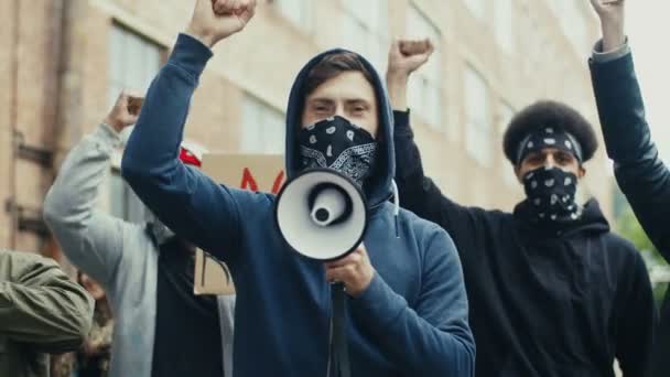 顔にフードとスカーフを着た白人の若いハンサムな男は、屋外の人権のための抗議でメガホンで叫んでいます。路上で抗議する人々のグループ。暴力に対する攻撃. — ストック動画
