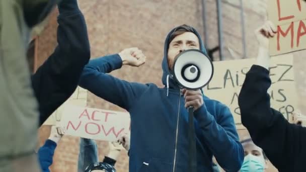 Biały, młody, przystojny mężczyzna w kapturze krzyczy megafonem na protest o prawa człowieka i wartości demokracji na zewnątrz. Facet w tłumie protestuje na ulicy. Uderzenie w przemoc. — Wideo stockowe