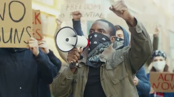Afroamerikan ung stilig man i halsduk i ansiktet som mask skriker i megafon mot protest för mänskliga rättigheter utomhus. En grupp människor protesterar på gatan. Strejk mot våld. — Stockvideo