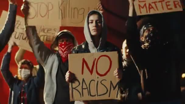 Mujer hermosa caucásica en la capucha de pie en la multitud de personas en la protesta callejera con cartel No hay racismo por la noche. Manifestación de Estados Unidos por la igualdad de derechos humanos. Manifestante femenina . — Vídeos de Stock