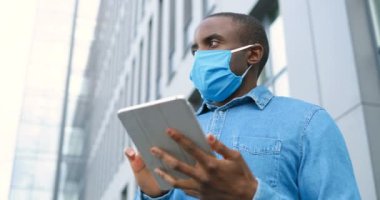 Tıbbi maskeli Afro-Amerikan bir adam sokakta duruyor ve tablet cihazına mesaj yolluyor. Solunum korumalı erkek dışarıda ve bilgisayara dokunuyor ya da kaydırıyor. Çevrimiçi tarama.