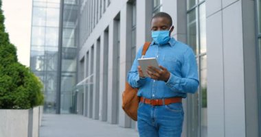 Tıbbi maskeli Afro-Amerikan bir adam sokakta duruyor ve tablet bilgisayarına mesaj yolluyor. Solunum korumalı bir erkek dışarıda yürüyor ve cihaza dokunuyor ya da kaydırıyor. Çevrimiçi tarama.
