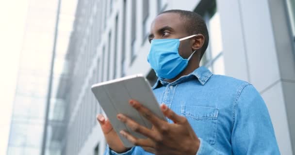 Afroamerykanin w masce medycznej stojący na ulicy i SMS-ujący na tablecie. Mężczyzna w ochronie dróg oddechowych jest na zewnątrz i stukanie lub przewijanie na komputerze. Przeglądanie online. — Wideo stockowe