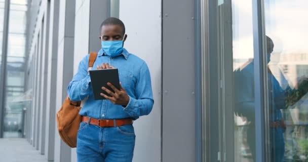 Hombre afroamericano con máscara médica de pie en la calle y mensajes de texto en la computadora tableta. Hombre en protección respiratoria caminando al aire libre y tocando o desplazándose en el dispositivo. Navegar en línea . — Vídeos de Stock