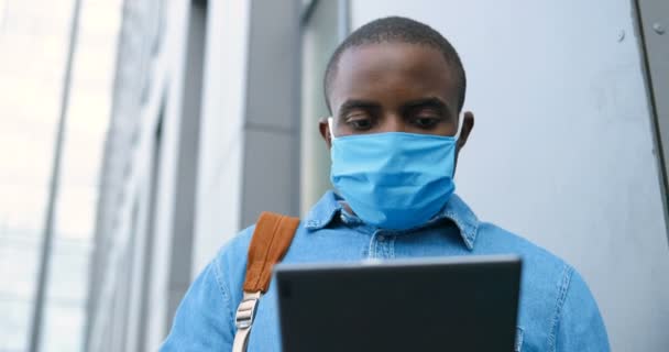 Homem afro-americano com máscara médica em pé na rua e mensagem de texto no dispositivo tablet. Masculino em proteção respiratória é ao ar livre e tocando ou rolando no computador. Navegação on-line . — Vídeo de Stock