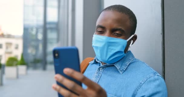 Αφροαμερικάνος με ιατρική μάσκα στέκεται στο δρόμο και βιντεοσκοπεί στο κινητό τηλέφωνο. Άνδρας στην αναπνευστική προστασία είναι σε εξωτερικούς χώρους, ενώ μιλάμε και να έχουν videochat από κάμερα στο smartphone — Αρχείο Βίντεο