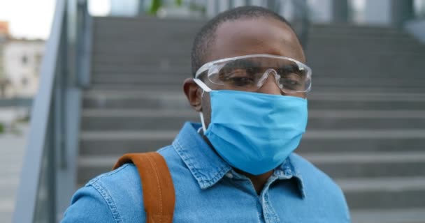 Közelkép a fiatal jóképű afro-amerikai férfi boldog arcáról orvosi maszkban és védőszemüvegben a szabadban. Portré felvétel vidám férfi visel légúti védelem az utcán. — Stock videók