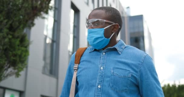 Porträtt av afroamerikan snygg ung man med ryggsäck, i medicinsk mask och glasögon står utomhus, vända ansikte mot kamera. Manlig fotgängare i andningsskydd. — Stockvideo