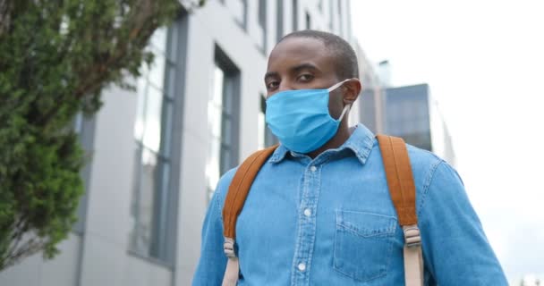 Porträtt av afroamerikan snygg ung man med ryggsäck och i medicinsk mask står utomhus, vända ansikte mot kamera och ser rakt. Manlig fotgängare i andningsskydd. — Stockvideo
