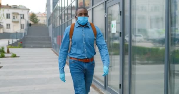 Молодой афроамериканец в медицинской маске ходит по улице и куда-то направляется. Мужской пешеход в дыхательной защите прогуливается под открытым небом. Красавчик прогуливается. Коронавирусная концепция . — стоковое видео