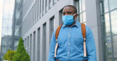 Tıbbi maskeli ve sırt çantalı Afro-Amerikan bir gencin portre fotoğrafı. Dışarıda dikiliyor, yüzünü kameraya çevirip dümdüz bakıyor. Erkek yaya, solunum koruması altında..