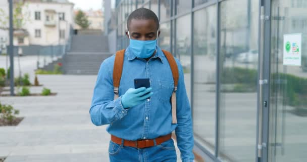 Afroamerikansk man i medicinsk mask strosar på gatan och sms: ar på mobilen. Man i andningsskydd promenader utomhus och knacka eller rulla på smartphone. Bläddrar på nätet. — Stockvideo