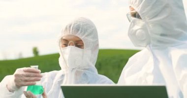 Close up of Caucasian female and male farmers researchers holding test tube with chemicals pesticides and laptop computer in green field. Biologists co-workers working and talking in wheat margin.