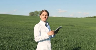 Caucasian beautiful female ecologist scientist in white gown walking in green field and holding tablet device. Woman researcher, biologist strolling in margin of harvest while using tablet computer.