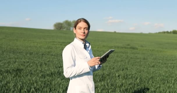 Caucasian beautiful female ecologist scientist in white gown walking in green field and holding tablet device. Woman researcher, biologist strolling in margin of harvest while using tablet computer. — ストック動画