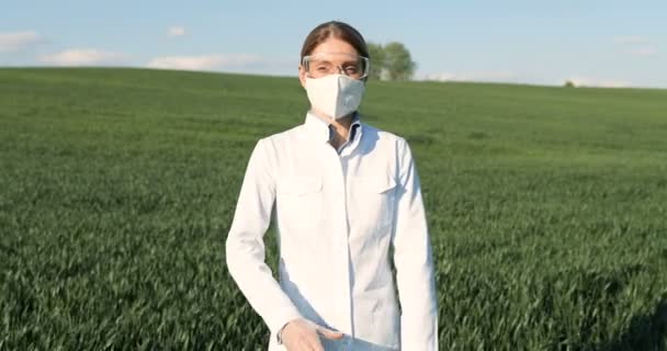 Portrait of Caucasian beautiful female ecologist scientist in white gown, mask and googles standing in green field and looking at camera with crossed hands. Woman researcher, biologist in margin. — Stock video